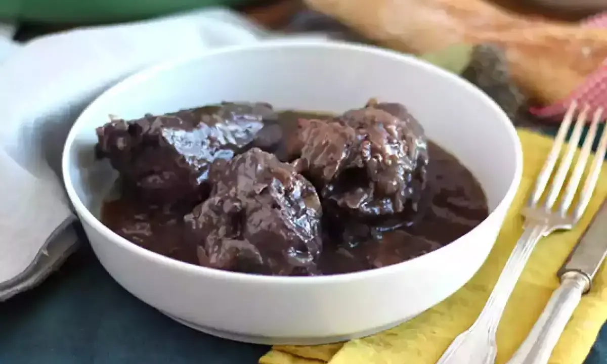 Un plat de coq au vin mijoté, servi dans un bol blanc, avec une fourchette et un couteau à côté.