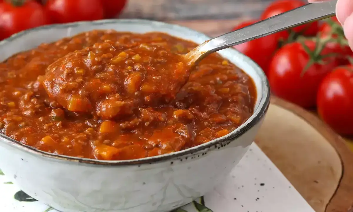 Une cuillère prélève une portion généreuse de sauce bolognaise maison, riche et savoureuse, dans un bol. Des tomates fraîches en arrière-plan.
