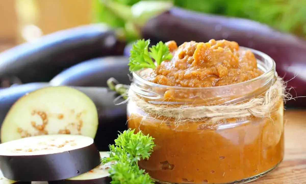 Un bocal de le vrai caviar d'aubergine, garni de persil frais, avec des aubergines entières et tranchées en arrière-plan.