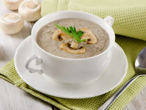 Un réconfortant velouté de champignon, garni de lamelles et d'une touche de persil, servi dans une tasse blanche sur une serviette verte.