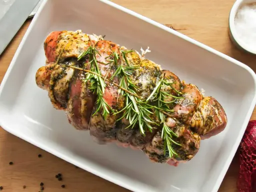 Un rôti de porc cru, assaisonné et garni de romarin frais, prêt à être cuit dans un plat blanc.
