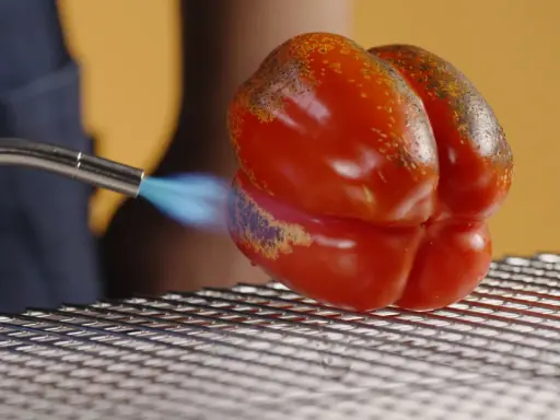 Un poivron rouge est rôti à la flamme sur une grille. La peau noircit par endroits, signe d'un poivron au four savoureux.