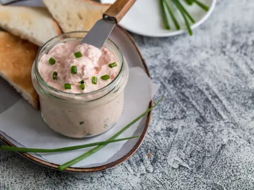 Un pot de rillettes de saumon crémeuses, garni de ciboule, prêt à être tartiné sur des toasts grillés.