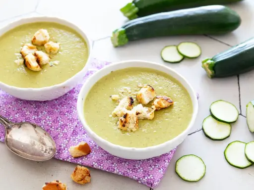 Deux bols de velouté de courgettes crémeux, garnis de croûtons dorés, avec des courgettes fraîches et des rondelles en arrière-plan.