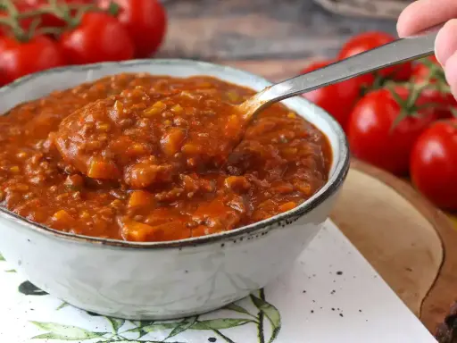 Une cuillère prélève une portion généreuse de sauce bolognaise maison, riche et savoureuse, dans un bol. Des tomates fraîches en arrière-plan.