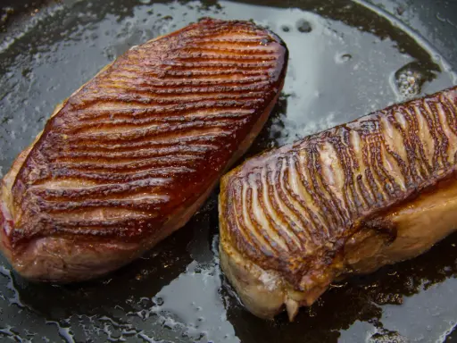 Deux magrets de canard au four, la peau grillée et dorée, baignant dans leur jus de cuisson sur une poêle.