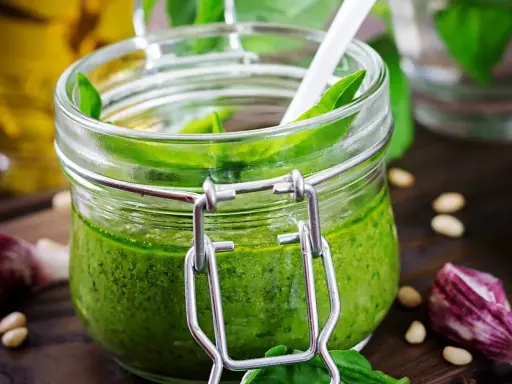 Un pot de pesto ail des ours, vert vif, avec une cuillère blanche, des pignons de pin et de l'ail sur un fond en bois.