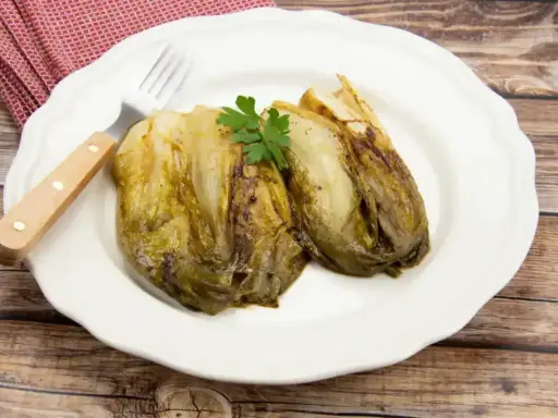 Deux endives caramélisées à la poêle, garnies de persil, sur une assiette blanche avec une serviette à carreaux rouges et une fourchette.