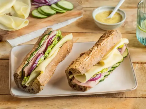 Deux sandwichs jambon fromage appétissants, garnis de concombre, oignon rouge et laitue, sur un plateau blanc.