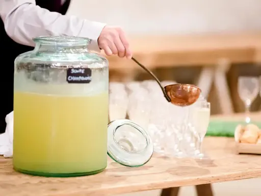 Une louche sert une soupe champenoise sans alcool dans un grand pichet en verre. Des verres à pied et des amuse-bouches sont disposés sur la table.