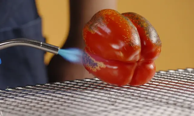 Un poivron rouge est rôti à la flamme sur une grille. La peau noircit par endroits, signe d'un poivron au four savoureux.