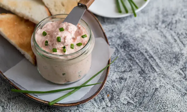 Un pot de rillettes de saumon crémeuses, garni de ciboule, prêt à être tartiné sur des toasts grillés.