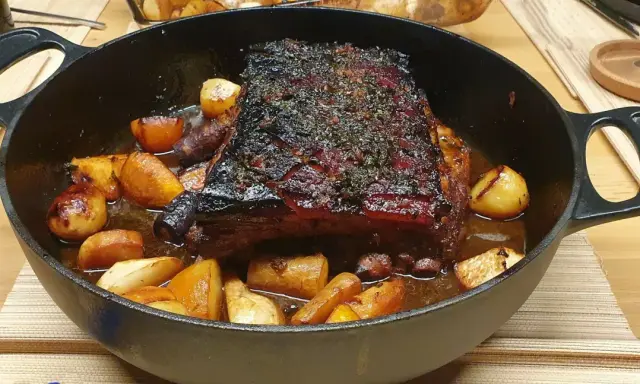 Poitrine de porc rôtie, croustillante et savoureuse, servie avec des pommes de terre et des carottes dans une cocotte en fonte.