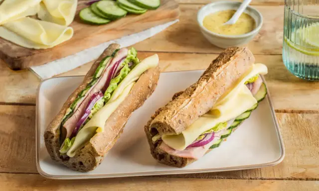 Deux sandwichs jambon fromage appétissants, garnis de concombre, oignon rouge et laitue, sur un plateau blanc.