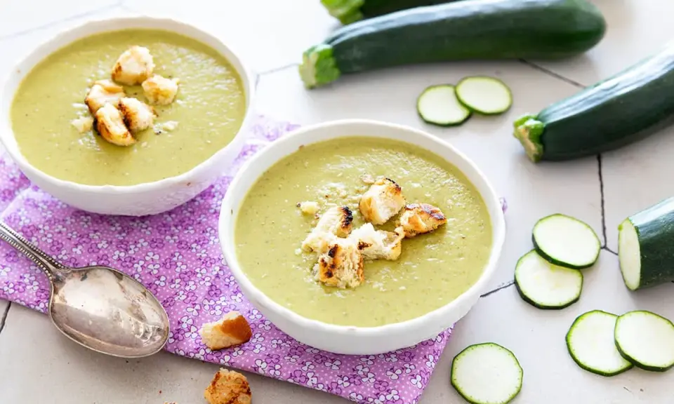 Deux bols de velouté de courgettes crémeux, garnis de croûtons dorés, avec des courgettes fraîches et des rondelles en arrière-plan.
