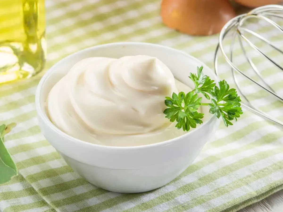 Un bol de mayonnaise onctueuse, garni de persil frais. Les ingrédients pour une recette mayonnaise parfaite : œufs, huile, et un fouet.