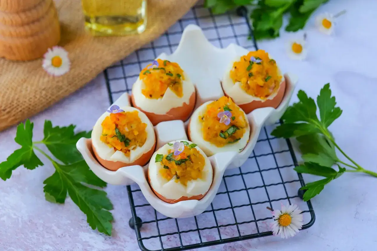 Cinq œufs mimosa garnis d'une purée orangée et d'herbes, décorés de petites fleurs violettes, présentés dans un plat blanc en forme d'œuf.