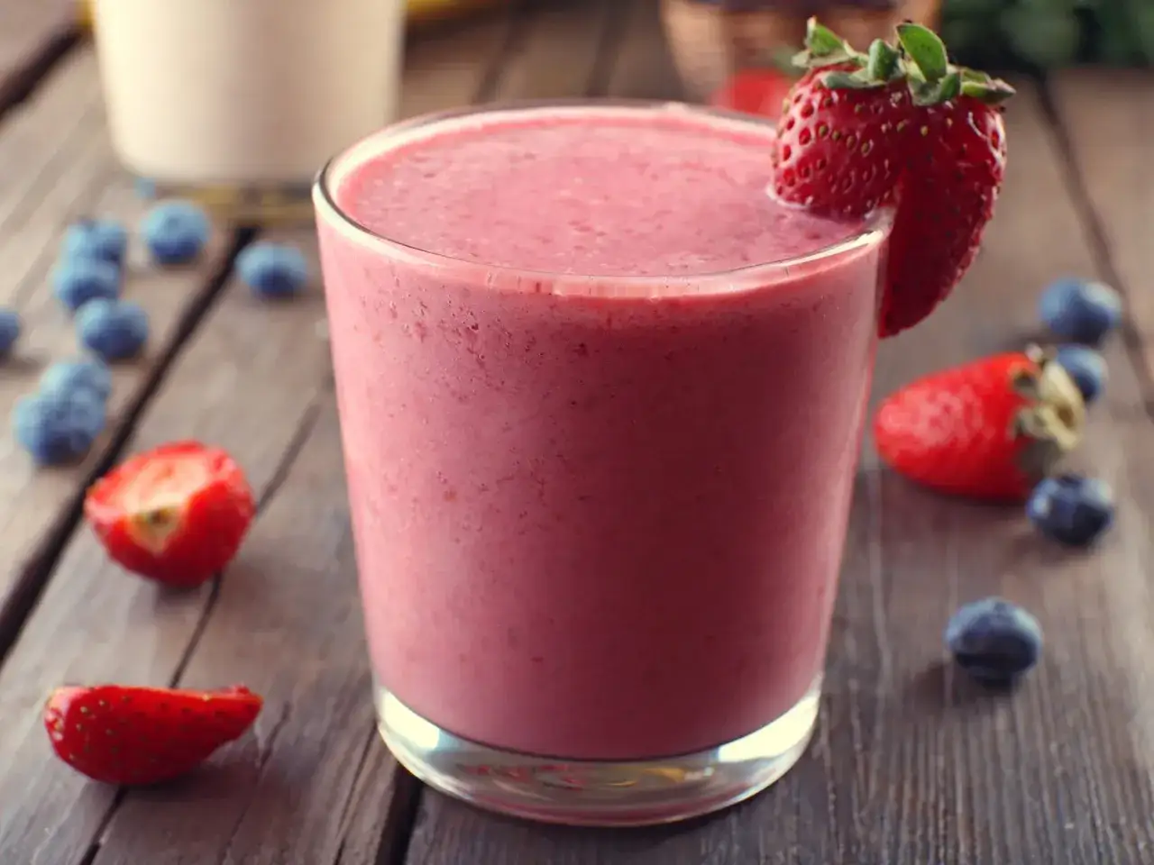 Un délicieux smoothie fraise banane, garni d'une fraise fraîche, entouré de fruits rouges sur une table en bois.