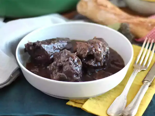Un plat de coq au vin mijoté, servi dans un bol blanc, avec une fourchette et un couteau à côté.
